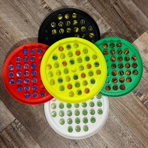 May include: Five circular trays with a white, yellow, red, green, and black background. Each tray has 49 small, clear glass marbles with different colored swirls inside.