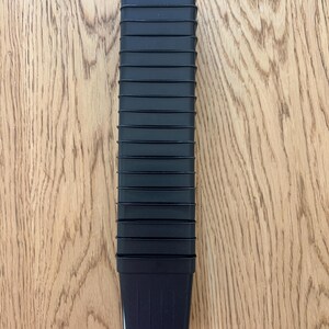 May include: A stack of black plastic plant pots, rectangular in shape, with a ridged design. The pots are empty and ready for use. The pots are sitting on a light brown wooden surface.