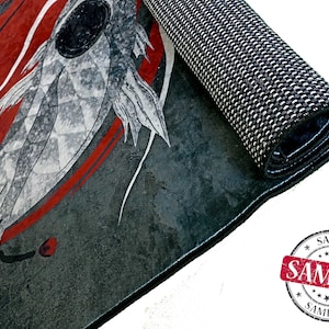Op de afbeelding: Een grijs en rood tapijt met een zwart-wit visontwerp. Het tapijt is opgerold en de rand van het tapijt is zichtbaar. Het tapijt heeft een antislip rug.