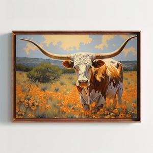 Puede incluir: Una impresión artística enmarcada que muestra una vaca longhorn con pelaje blanco y marrón de pie en un campo de flores de color naranja vibrante. El fondo muestra un paisaje ondulado bajo un cielo pálido con nubes suaves, en un marco de madera.