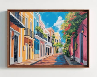 May include: A framed painting depicts a colorful street scene. Buildings in shades of orange, blue, and pink line the street, with balconies and windows. A vibrant blue sky with clouds is visible above. Red flowers and green vines cascade down the side of a building.