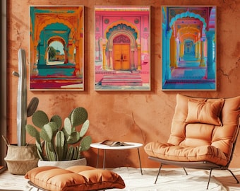Op de afbeelding: Drie rechthoekige wandkunstprints met levendige architecturale scènes met boogdeuren en zuilengangen in tinten roze, oranje en blauw, op een rij weergegeven op een getextureerde terracotta-kleurige muur boven een cactus en stoel.