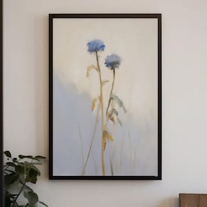 Puede incluir: Una pintura de acuarela enmarcada con dos flores azules con tallos marrones sobre un fondo suave y claro. La obra de arte está encerrada en un marco negro, creando una estética minimalista. La pintura evoca una sensación de calma y belleza natural.