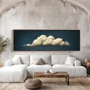 May include: A large rectangular framed artwork featuring a fluffy white cloud against a dark blue background. The artwork hangs above a white sectional sofa in a neutral-toned living room. The room includes a woven coffee table and decorative vases.