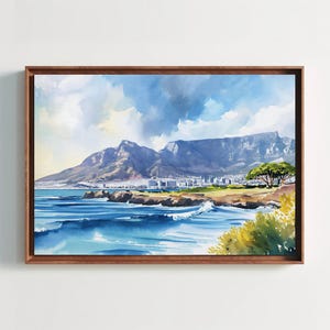 May include: A framed watercolor painting depicting a coastal landscape with blue ocean waves crashing against a rocky shoreline. In the background, a large mountain range rises under a cloudy sky, with a city visible along the coast.