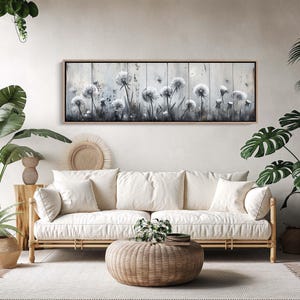 Puede incluir: Una obra de arte rectangular enmarcada presenta una pintura de acuarela de dientes de león blancos sobre un fondo de madera gris claro y desgastado. La obra de arte se exhibe sobre un sofá crema con cojines a juego, creando una estética serena y natural.