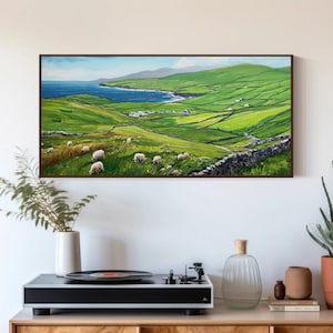 May include: A rectangular landscape painting featuring a vibrant green hillside with grazing white sheep, stone walls, and a distant blue ocean under a light sky. The artwork is framed in dark wood and displayed on a wall above a record player.