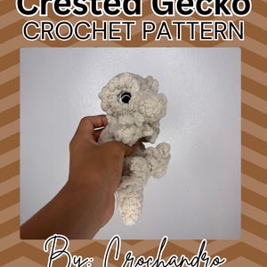 Op de afbeelding: Een gehaakte gekko in een lichtbeige kleur, vastgehouden in een hand. De gekko heeft grote, ronde ogen en een getextureerd uiterlijk. Tekst op de afbeelding luidt "Crested Gecko CROCHET PATTERN" en "By: Crochandra".