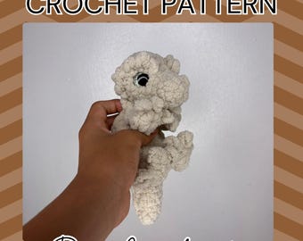 May include: A crocheted gecko in a light beige colour, held in a hand. The gecko has large, round eyes and a textured appearance. Text on the image reads "Crested Gecko CROCHET PATTERN" and "By: Crochandra".