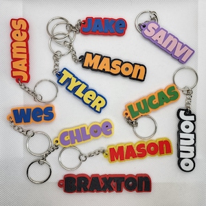May include: A selection of name keyrings in various colours. Each keyring displays a different name in bold, bright colours such as red, blue, orange, green, and purple. The keyrings are attached to silver key rings.