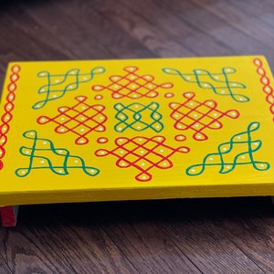 Peut inclure: Un plateau en bois jaune avec un motif géométrique rouge, vert et blanc peint sur le dessus. Le plateau a un bord surélevé et quatre petites pattes.