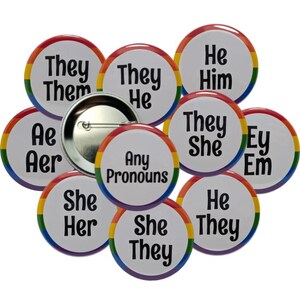 May include: A collection of pronoun buttons with rainbow borders. The buttons display pronouns such as "They/Them", "He/Him", "She/Her", and "Any Pronouns". A silver pinback button is in the center.
