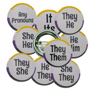 May include: A collection of round pronoun buttons with white backgrounds, yellow and purple borders, and various pronouns printed in black. The buttons include "They/Them", "He/Him", "She/Her", "It/Its", and "Any Pronouns".