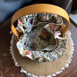 May include: A round, woven basket with a floral fabric lining. The basket has a wooden handle and is sitting on a burlap base with a white lace trim.