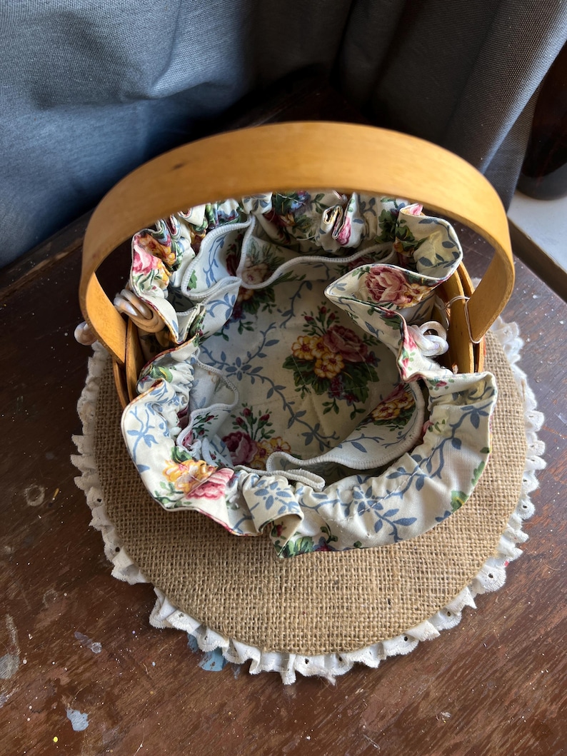 May include: A round, woven basket with a floral fabric lining. The basket has a wooden handle and is sitting on a burlap base with a white lace trim.