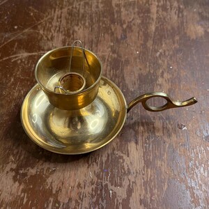 May include: A vintage brass candle holder with a circular base and a small handle. The candle holder has a cup-shaped design to hold the candle and a small handle for easy carrying. The brass has a warm, golden hue.