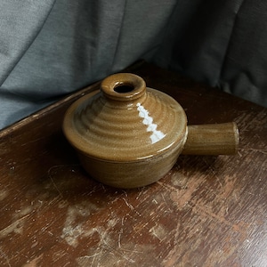 Puede incluir: Una olla de cerámica marrón con asa y una pequeña abertura en la parte superior. La olla tiene un esmalte moteado y una superficie ligeramente texturizada.