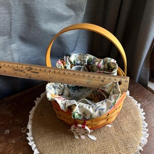 May include: A yellow and orange woven basket with a floral fabric liner. The basket has a wooden handle and a small charm attached to it. The basket is sitting on a brown burlap surface with white lace trim.