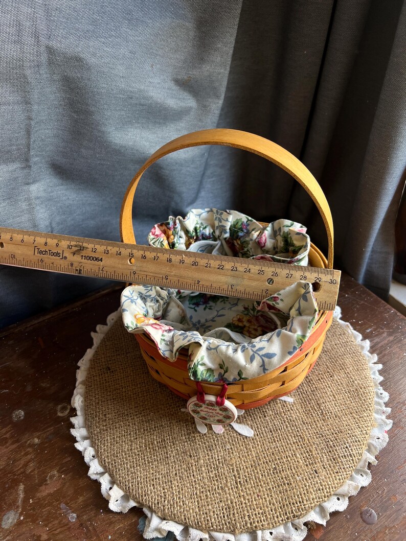 May include: A yellow and orange woven basket with a floral fabric liner. The basket has a wooden handle and a small charm attached to it. The basket is sitting on a brown burlap surface with white lace trim.
