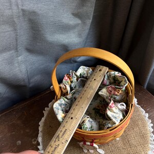 May include: A small, round, wooden basket with a fabric liner featuring a floral pattern. The basket has a wooden handle and is sitting on a burlap surface. A ruler is placed on top of the basket for size reference.