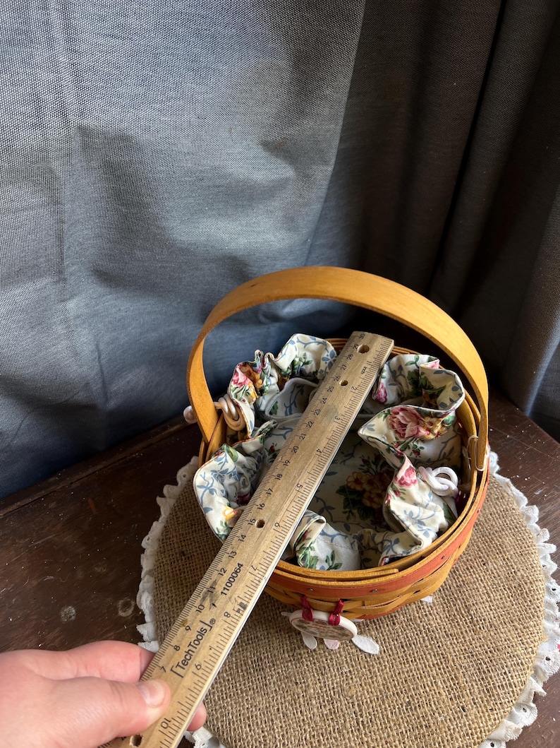 May include: A small, round, wooden basket with a fabric liner featuring a floral pattern. The basket has a wooden handle and is sitting on a burlap surface. A ruler is placed on top of the basket for size reference.