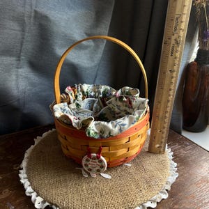 May include: A small, woven basket with a red and orange base and a natural wood handle. The basket is lined with a floral fabric and has a red ribbon attached to a round, ceramic ornament with a floral design and the text "Happy Birthday to You". The basket is sitting on a round, burlap placemat with a white lace trim.