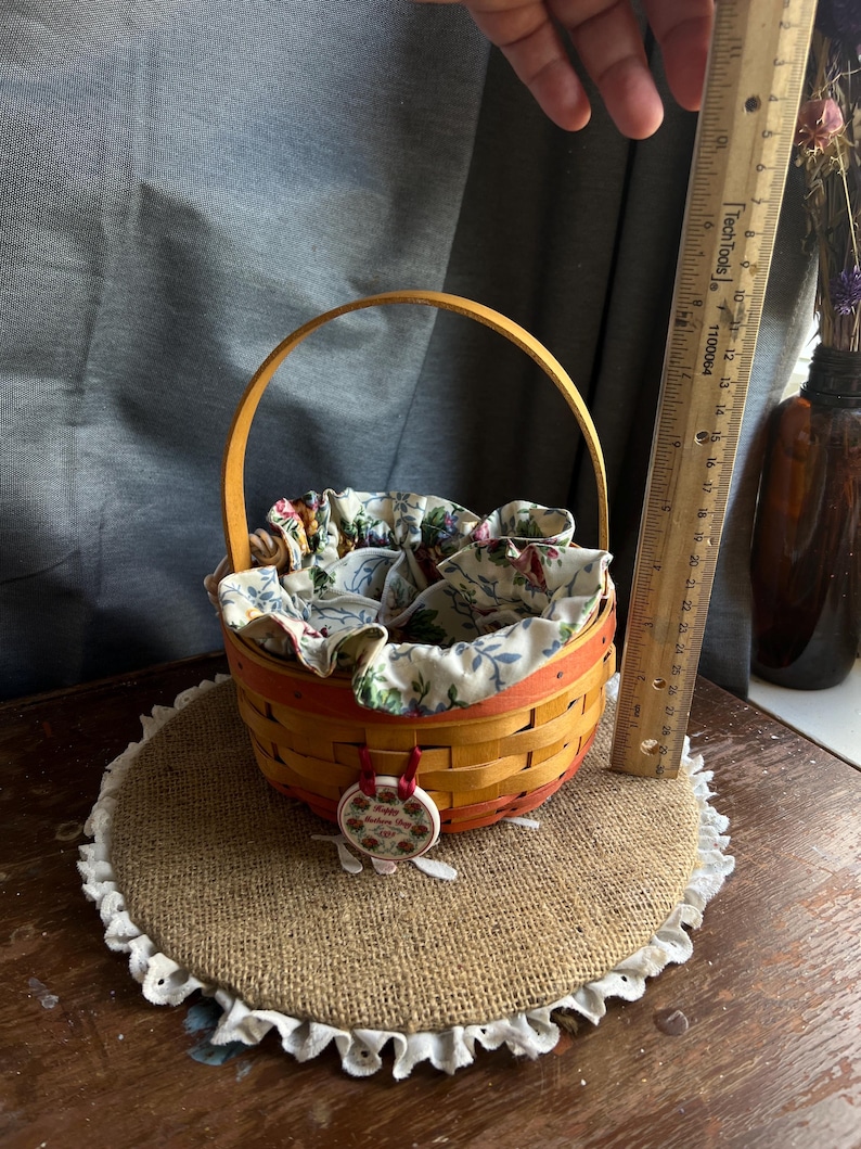 May include: A small, woven basket with a red and orange base and a natural wood handle. The basket is lined with a floral fabric and has a red ribbon attached to a round, ceramic ornament with a floral design and the text "Happy Birthday to You". The basket is sitting on a round, burlap placemat with a white lace trim.