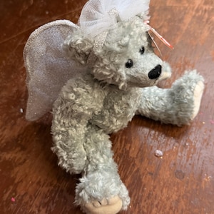 May include: A gray teddy bear with a white tulle bow and silver glitter wings. The bear has a black nose and small black eyes. The bear is lying on a wooden surface.