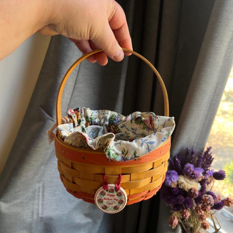 May include: A small, woven basket with a red and orange trim. The basket is lined with a floral fabric and has a tag that reads "Happy Mother's Day".
