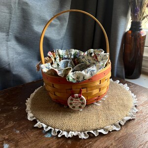 May include: A small, woven basket with a wooden handle and a red and white floral lining. The basket is sitting on a burlap mat with a white lace trim. A red and white ceramic tag with the text "Happy Mother's Day" is attached to the handle.
