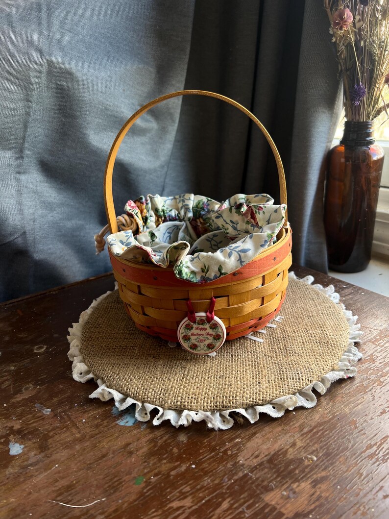 May include: A small, woven basket with a wooden handle and a red and white floral lining. The basket is sitting on a burlap mat with a white lace trim. A red and white ceramic tag with the text "Happy Mother's Day" is attached to the handle.