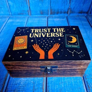 May include: A wooden box with a dark blue lid featuring the text "TRUST THE UNIVERSE". The lid has illustrations of the sun, moon, hands, and stars. The box is rectangular with a metal clasp.