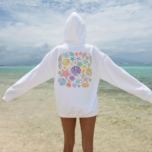 May include: White hooded sweatshirt with a colorful seashell and starfish design on the back. The design includes various colors such as purple, orange, blue, and yellow. The sweatshirt is a casual, comfortable garment.