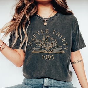 May include: A charcoal grey short-sleeve t-shirt featuring a gold-colored graphic of an open book with wildflowers emerging from the pages. The text "CHAPTER THIRTY" is arched above the book, and the number "1995" is printed below the graphic.