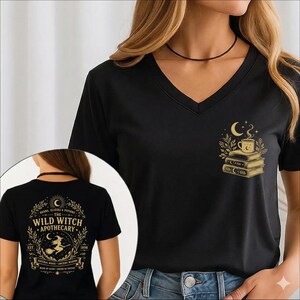 May include: A black V-neck t-shirt featuring a gold graphic on the chest of a steaming mug atop a stack of books. The back displays a gold circular emblem with the text "The Wild Witch Apothecary" and a witch flying on a broomstick.