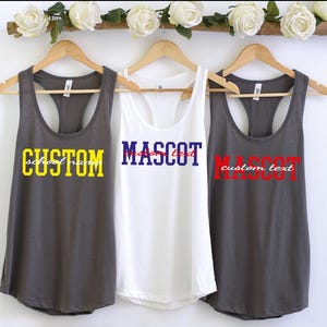 May include: Three racerback tank tops hanging on wooden hangers. The left is dark gray with yellow text reading CUSTOM and white text reading school name. The center is white with blue text reading MASCOT and red text reading custom text. The right is dark gray with red text reading MASCOT and white text reading custom text.