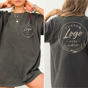 May include: Dark gray t-shirt with a round graphic on the front and back. The graphic features a mountain scene with the words "CUSTOM Logo - HERE" in a circular design. The shirt has short sleeves and a relaxed fit.