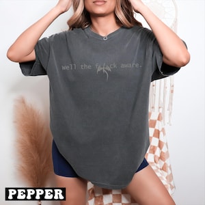 May include: Dark grey t-shirt with the text "well the f*ck aware" and a dragon graphic. The shirt is a relaxed fit and has short sleeves. The word "PEPPER" is printed in a white box at the bottom.