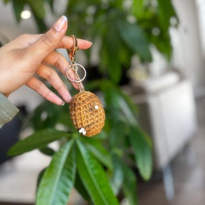 Pode incluir: Um chaveiro de crochê marrom feito à mão com fecho e argola dourados. O chaveiro tem um formato oval pequeno e é adornado com pequenas contas brancas. O fundo apresenta folhagem verde.