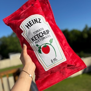 Pode incluir: Uma almofada de pelúcia em forma de saqueta de ketchup de tomate Heinz vermelha. A frente apresenta um rótulo branco com o texto HEINZ TOMATO KETCHUP AUX TOMATES, um gráfico de um tomate vermelho num ramo verde e o texto 8 mL em tinta preta.