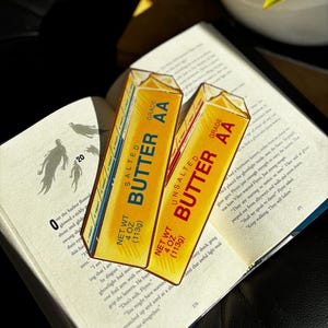 May include: Two butter-shaped bookmarks rest on an open book. One is labeled "Salted Butter" and the other "Unsalted Butter." Both have a yellow and white design with the text "Grade AA" and "Net Wt. 113g." The book is open to page 270.