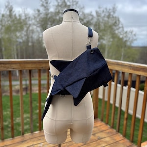 May include: A dark blue denim crossbody bag with a geometric, angular shape featuring a single shoulder strap attached with silver-toned metal hardware. The bag has a structured, folded design with visible dark buttons on the front flap surface.