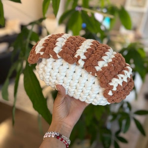 Puede incluir: Una almohada de sushi hecha a crochet en forma de nigiri con hilo blanco y marrón.