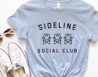 Op de afbeelding: Lichtblauw T-shirt met zwarte tekst die "Sideline Social Club" luidt en een afbeelding van drie regisseursstoelen.