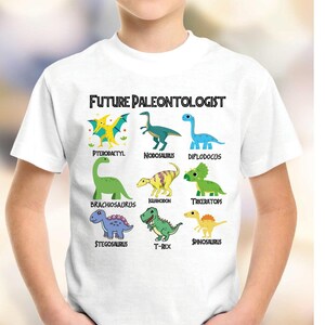 May include: White t-shirt featuring a colorful dinosaur graphic with the text "FUTURE PALEONTOLOGIST." The design includes illustrations of various dinosaurs such as Pterodactyl, Brachiosaurus, and T-Rex.