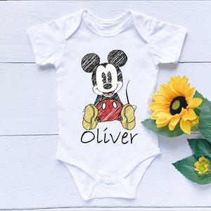 May include: White baby onesie featuring a hand-drawn style Mickey Mouse graphic. The design includes black ears, a red belly, yellow shoes, and the name "Oliver" in black script. A yellow sunflower with green leaves is next to the onesie.