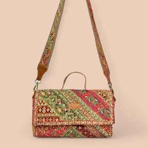 Può includere: Borsa ricamata rettangolare con tracolla. La borsa presenta un vivace mix di ricami rossi, verdi e dorati. La tracolla è marrone con cuciture decorative. La borsa ha un piccolo manico e una chiusura a patta. La parola "PRASHU" è visibile.