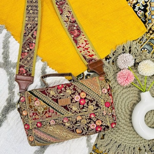 Può includere: Borsa a tracolla ricamata con cinturino decorativo. La borsa presenta un design patchwork con motivi floreali e geometrici nei toni del bordeaux, oro e rosa. Il cinturino è adornato da ricami simili e dettagli in pelle.