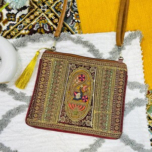 Può includere: Borsa a tracolla ricamata con cinturino in pelle marrone e nappa dorata. La borsa presenta un motivo paisley centrale con motivi floreali e di uccelli colorati, circondato da intricati abbellimenti dorati e verdi. La borsa è rettangolare e ha un bordo rosso.