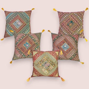 May include: Five decorative throw pillows with intricate embroidered patterns in shades of red, green, and gold. Each pillow features a central diamond design and is adorned with a yellow tassel at each corner. The pillows are arranged on a light pink background.